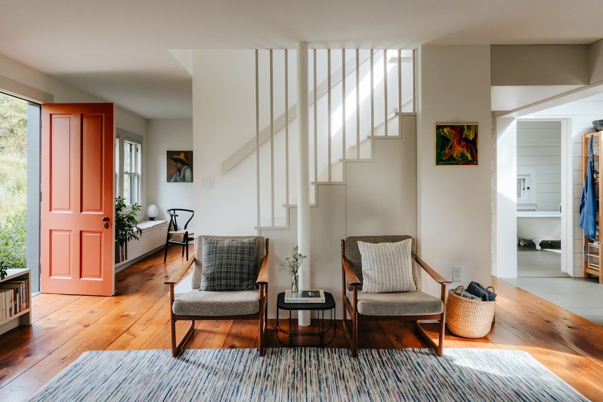 A bright double-height living room with a sculptural custom staircase rising beside a large window wall, featuring floating timber treads, slim steel supports, and a glass balustrade that keeps the space open and light