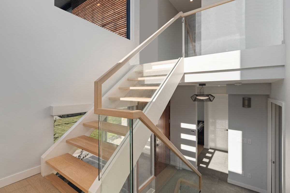 A split scene showing an indoor staircase with warm timber treads and glass balustrades on one side, and an outdoor staircase on the other side made of galvanised steel with concrete or stone steps, wet surfaces, and surrounding garden foliage under bright sun and stormy sky conditions.